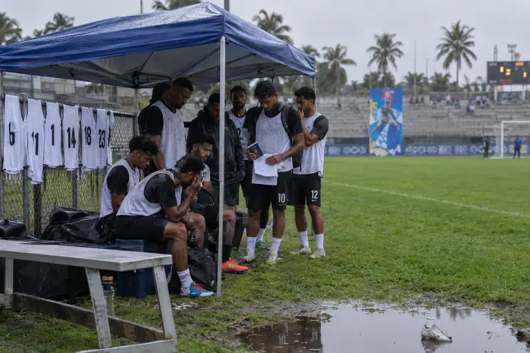 Players wait under a tarp on the training pitch in Suva after late eligibility rulings left squads short of registered players.